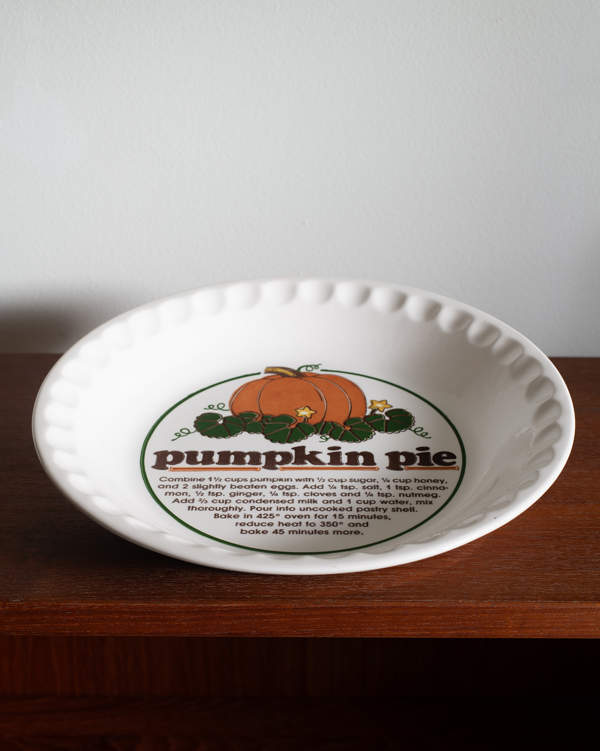A white ceramic pumpkin pie dish with a scalloped rim and a printed pumpkin pie graphic, sitting on a dark wooden surface against a light gray wall.