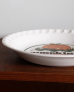 A white ceramic pie dish with a scalloped rim, featuring a printed autumn design with a pumpkin, green leaves, and the word 'pumpkin' on a dark wooden surface against a light gray wall.