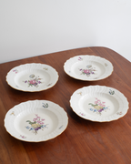 Four white ceramic plates with scalloped edges and delicate floral prints, arranged on a wooden table against a plain light background.
