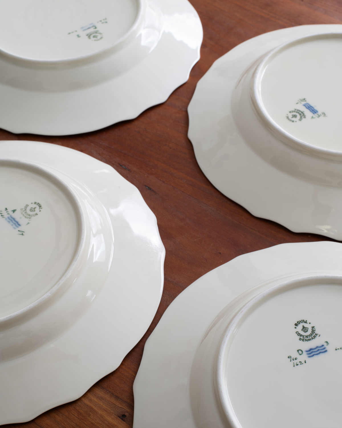 Four white ceramic plates with scalloped edges arranged on a brown wooden table, showing the printed maker's marks on the bottoms.