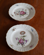 Two vintage white porcelain plates with gold trim and floral designs, displayed on a dark wooden table.