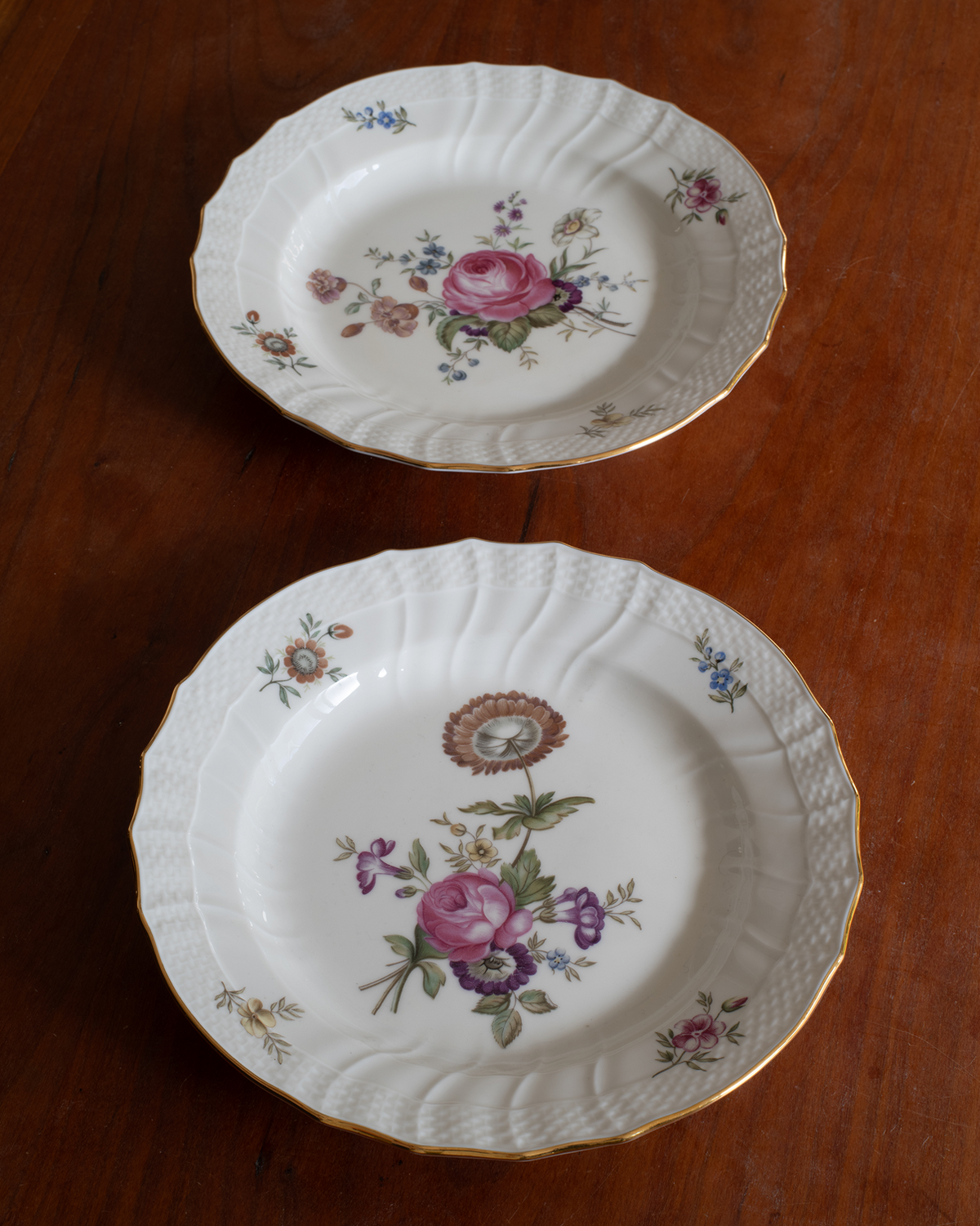 Two vintage white porcelain plates with gold trim and floral designs, displayed on a dark wooden table.