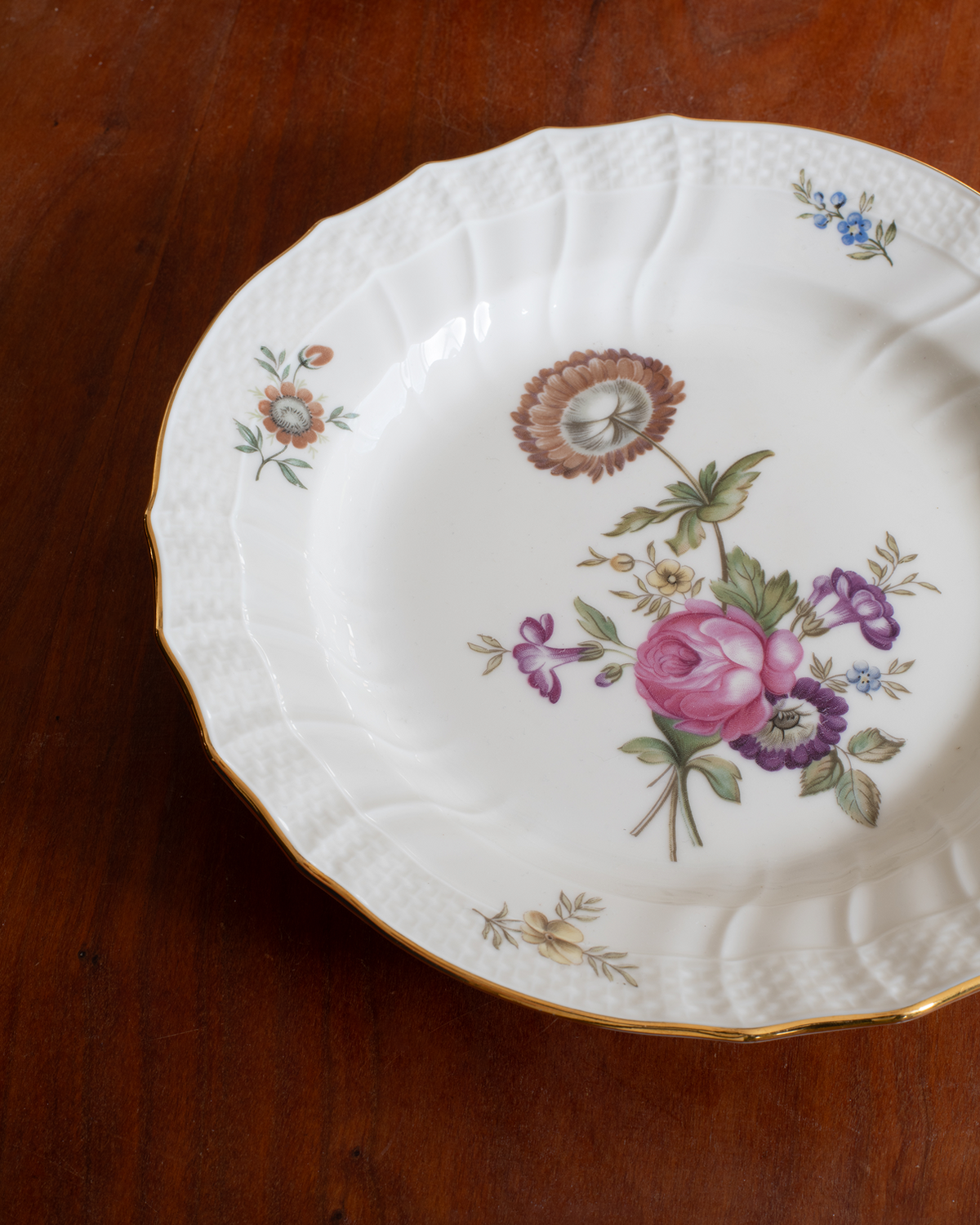 A white porcelain plate with a scalloped rim, gold trim, and delicate floral illustrations, photographed on a dark wooden tabletop.