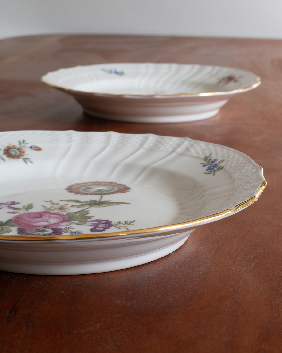 Two white porcelain plates with gold trim and floral prints resting on a wooden table.