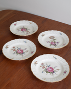 Four white porcelain plates with scalloped edges, decorated with pink rose floral motifs and gold trim, arranged on a wooden table.