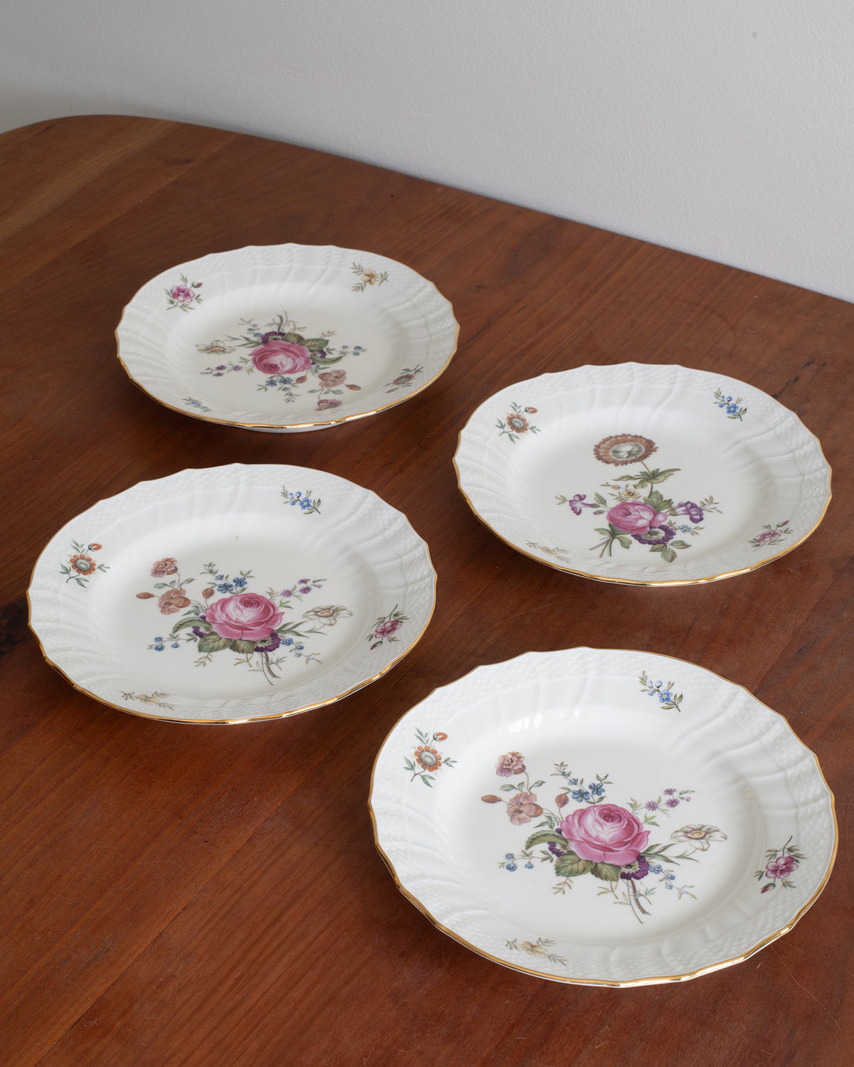 Four white porcelain plates with scalloped edges, decorated with pink rose floral motifs and gold trim, arranged on a wooden table.