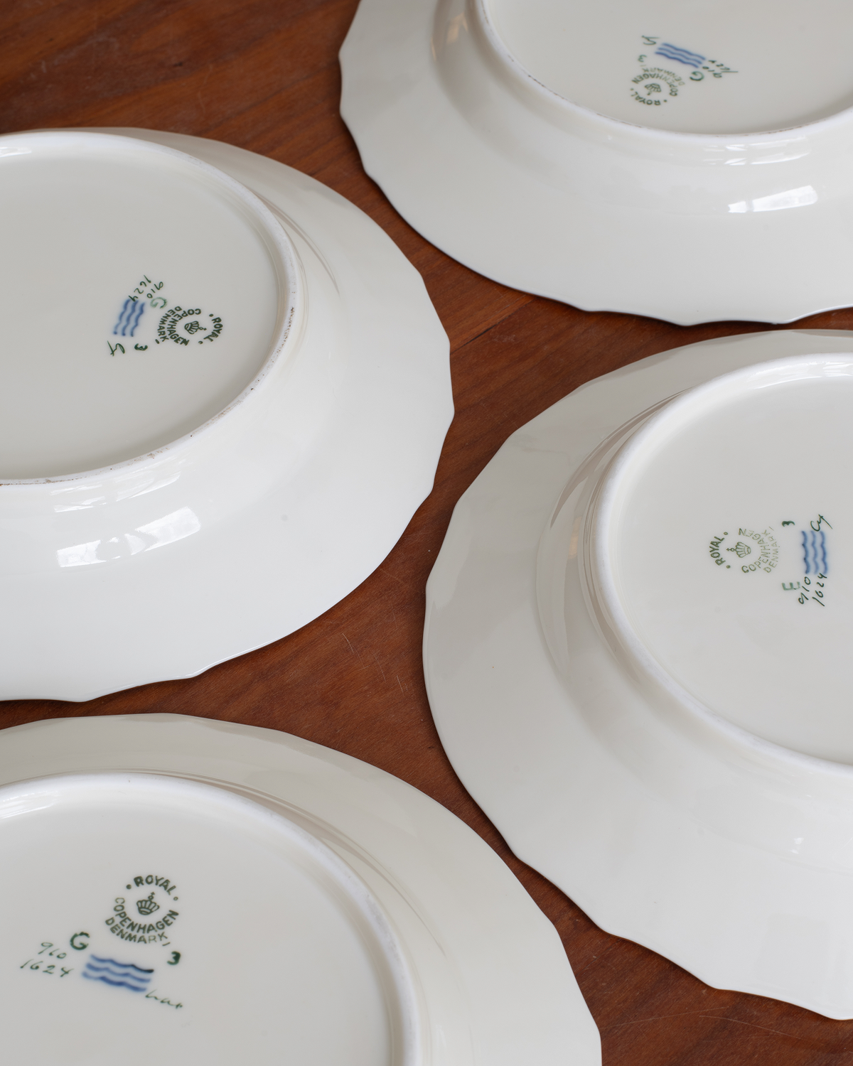 Four white ceramic plates arranged on a brown wooden surface, showing the inside bottoms with blue and green printed maker marks.