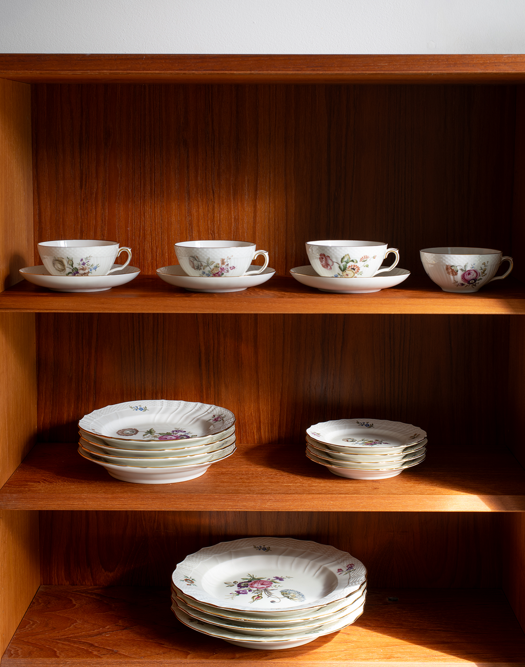 A set of white porcelain tableware with delicate floral prints, neatly arranged on wooden shelves. The collection includes teacups with saucers, plates, and bowls, showcasing a classic vintage pattern.