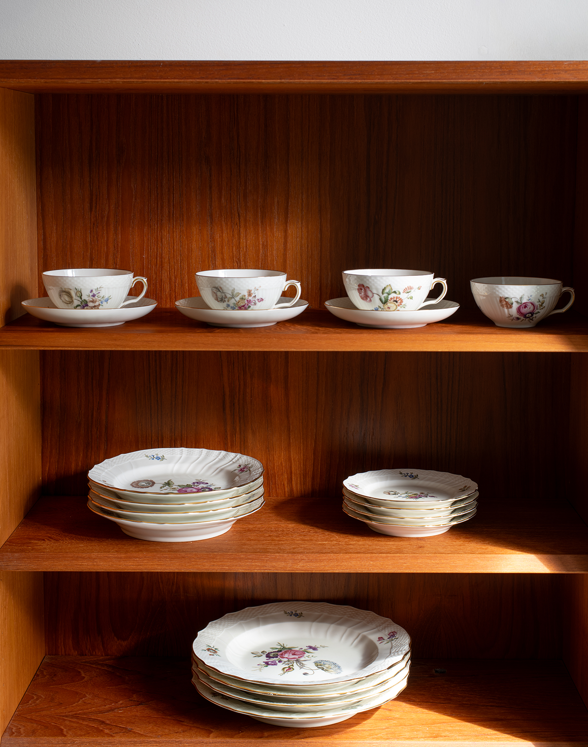 A set of white porcelain tableware with delicate floral prints, neatly arranged on wooden shelves. The collection includes teacups with saucers, plates, and bowls, showcasing a classic vintage pattern.