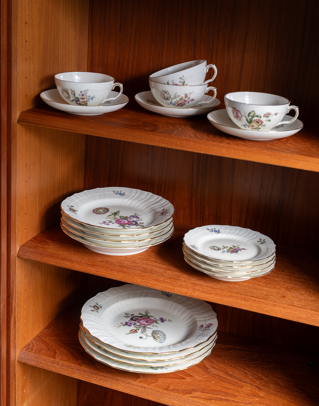 A set of white porcelain tea and coffee tableware with delicate pink, purple, and green floral patterns, displayed on three wooden shelves.