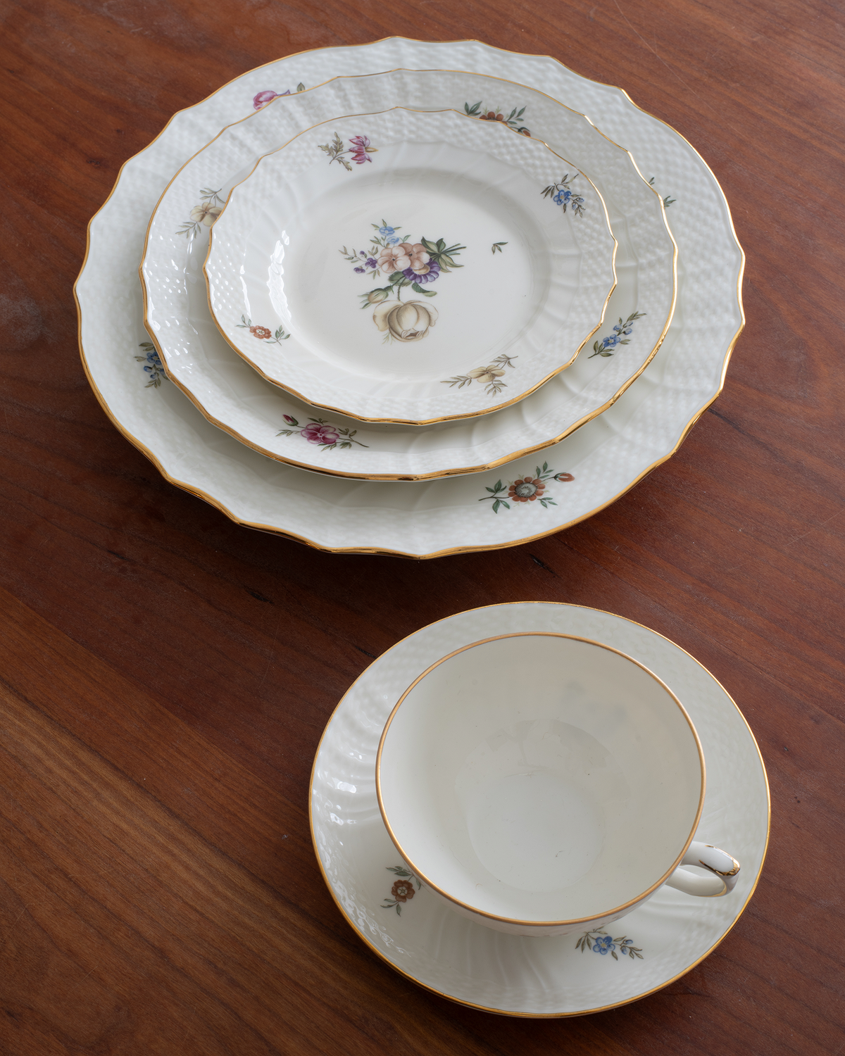 A set of white porcelain tableware with gold trim and delicate floral prints, arranged on a dark wooden table. The set includes a large platter, two dinner plates, a teacup on a saucer, and a smaller plate.