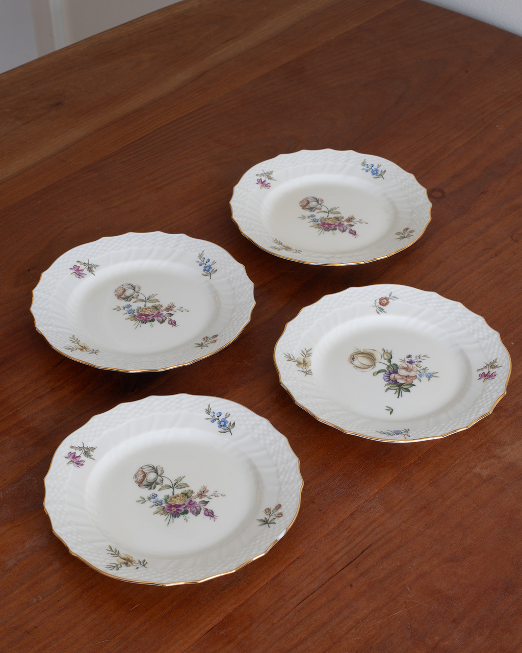 Four white porcelain plates with gold trim and floral prints, arranged on a wooden table.