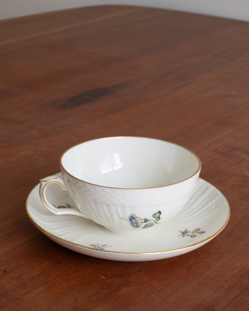 A white porcelain teacup and saucer set with delicate gold trim and small blue floral details, placed on a dark wooden table.