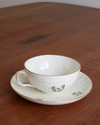 A white porcelain teacup and saucer set with delicate gold trim and small blue floral details, placed on a dark wooden table.