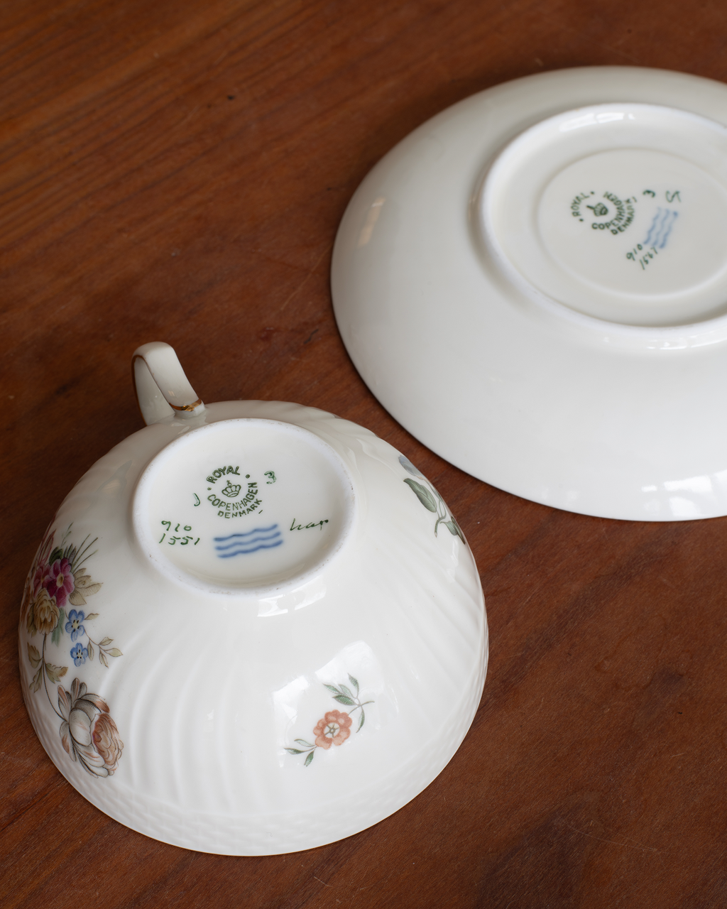 A white porcelain teacup and saucer set on a dark brown wooden table, showing the floral underglaze decoration and maker's marks.