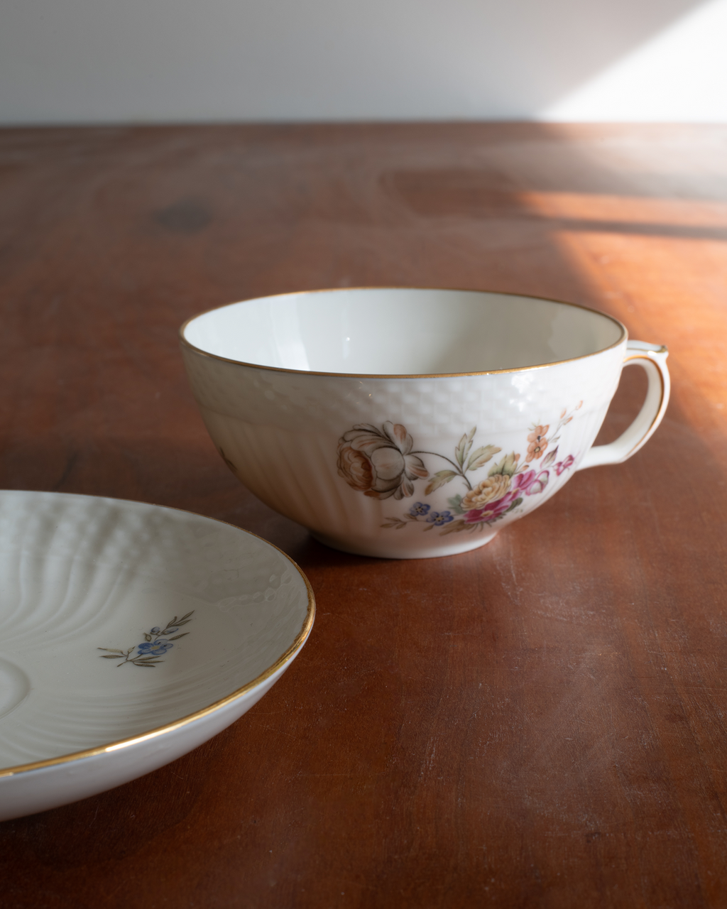 A white porcelain teacup with gold trim and a delicate floral pattern, shown alongside a matching saucer on a warm wooden tabletop with soft natural light.