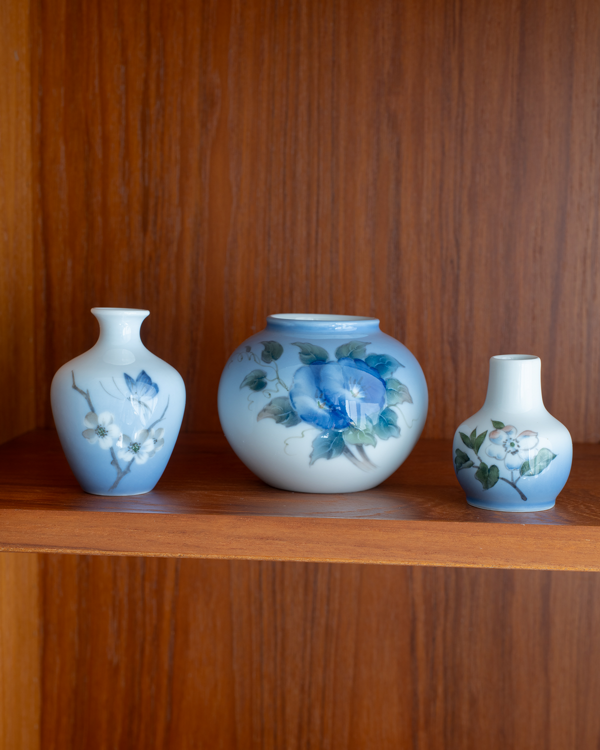Three blue and white ceramic vases with floral hand-painted designs displayed on a wooden shelf against a wooden wall.