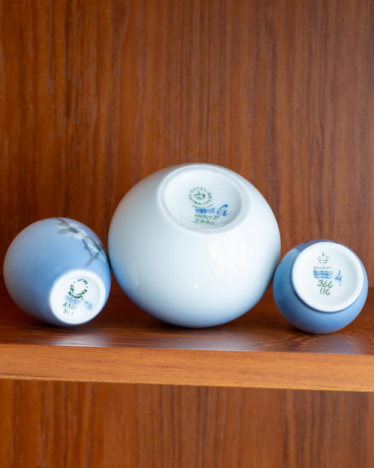 Three small glossy ceramic cups or saucers displayed upside down on a wooden shelf, with one larger white cup in the center and two smaller blue cups on either side, photographed against a wooden background.