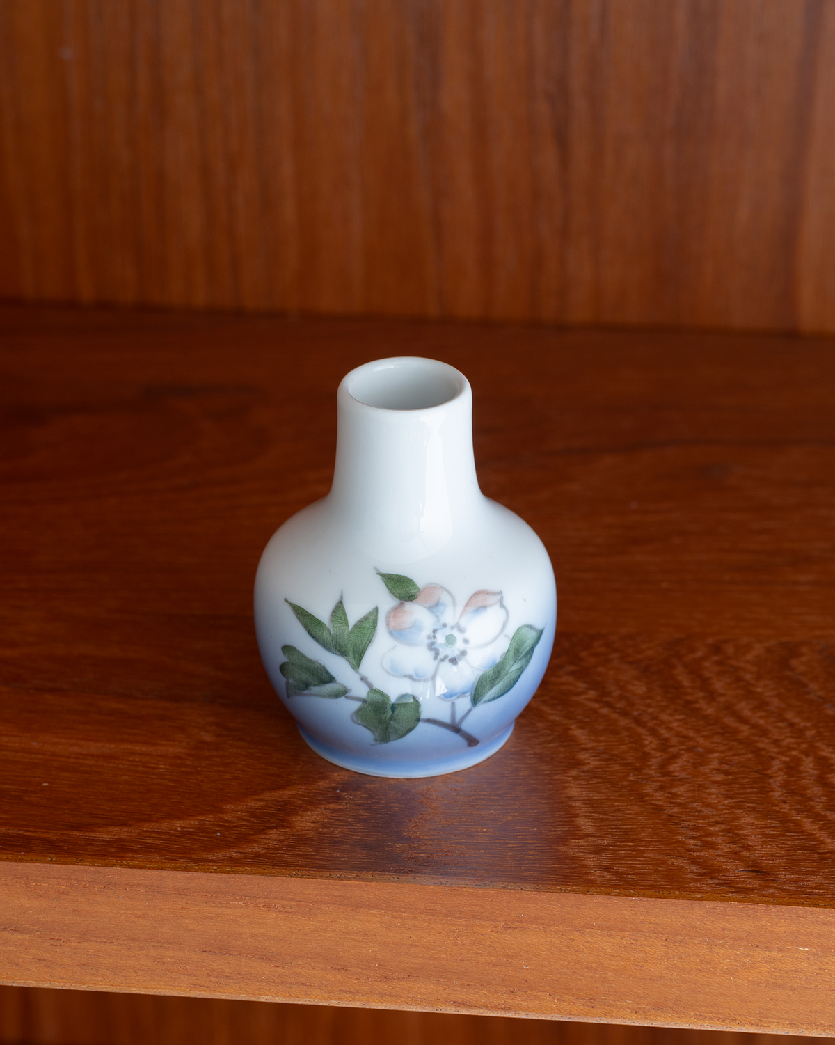A small ceramic bud vase with a white and blue glazed body, decorated with a hand-painted white flower and green leaves, sitting on a polished wooden surface.