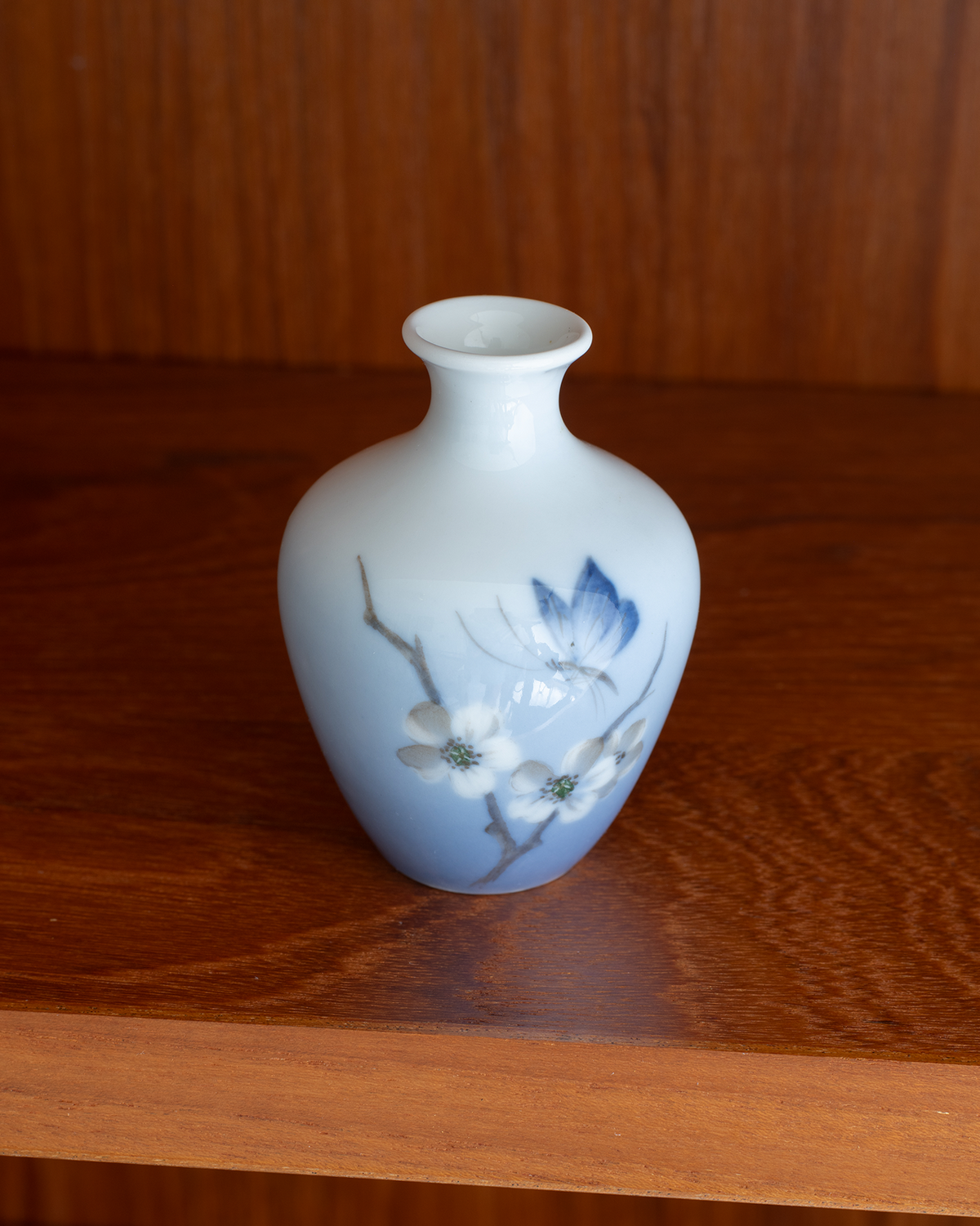 A small ceramic vase with a pale blue to white gradient body, decorated with white blossoms and blue leaves, sitting on a wooden shelf.