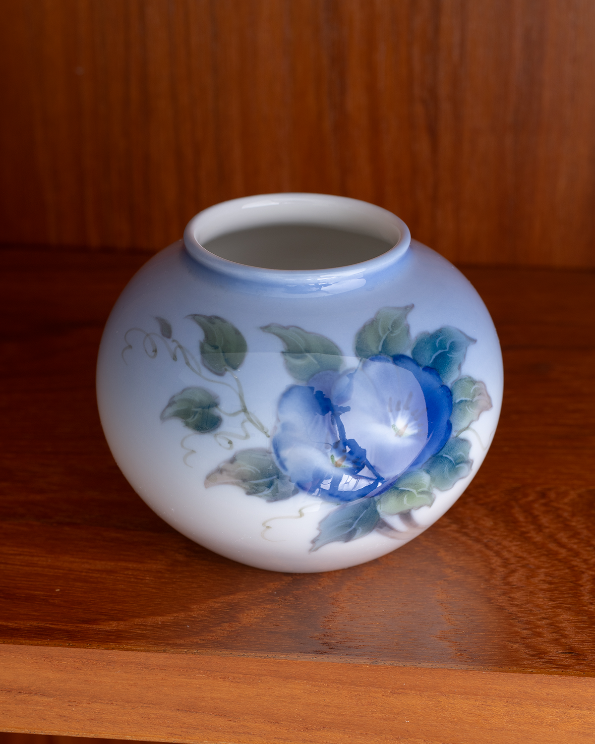 A small round ceramic vase with a glossy white glaze, decorated with blue and green floral motifs resembling morning flowers, displayed on a wooden shelf.