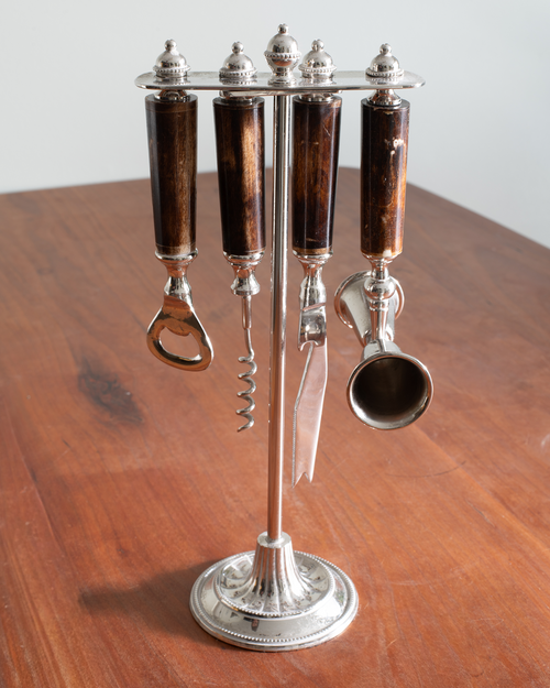 A vintage six-piece wine and liquor accessory set on a silver stand, featuring dark brown wooden handles and silver-toned metal tools including a corkscrew, bottle opener, scissors, and a small scoop, displayed on a wooden table.