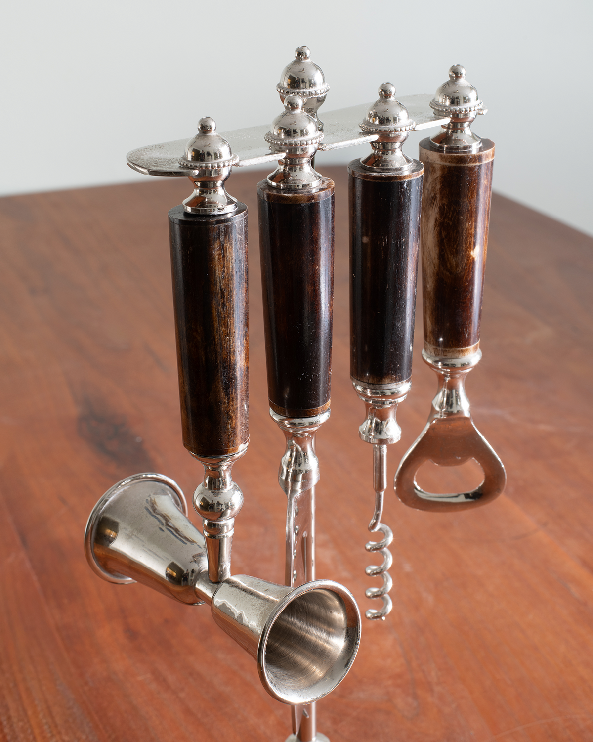 A set of four bar tools with dark brown wooden handles and polished silver-toned metal heads, including a corkscrew, bottle opener, and two small metal accessories, displayed on a wooden surface.