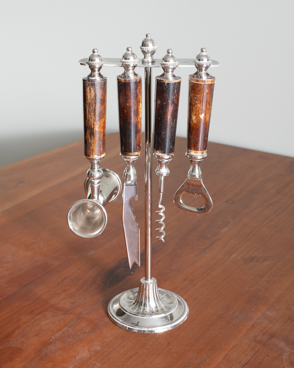 A vintage four-piece wine accessory set displayed on a wooden table. The set includes four bottles with dark wood handles, a corkscrew, a wine opener, a small spoon, and a knife, all mounted on a polished silver-tone metal stand.