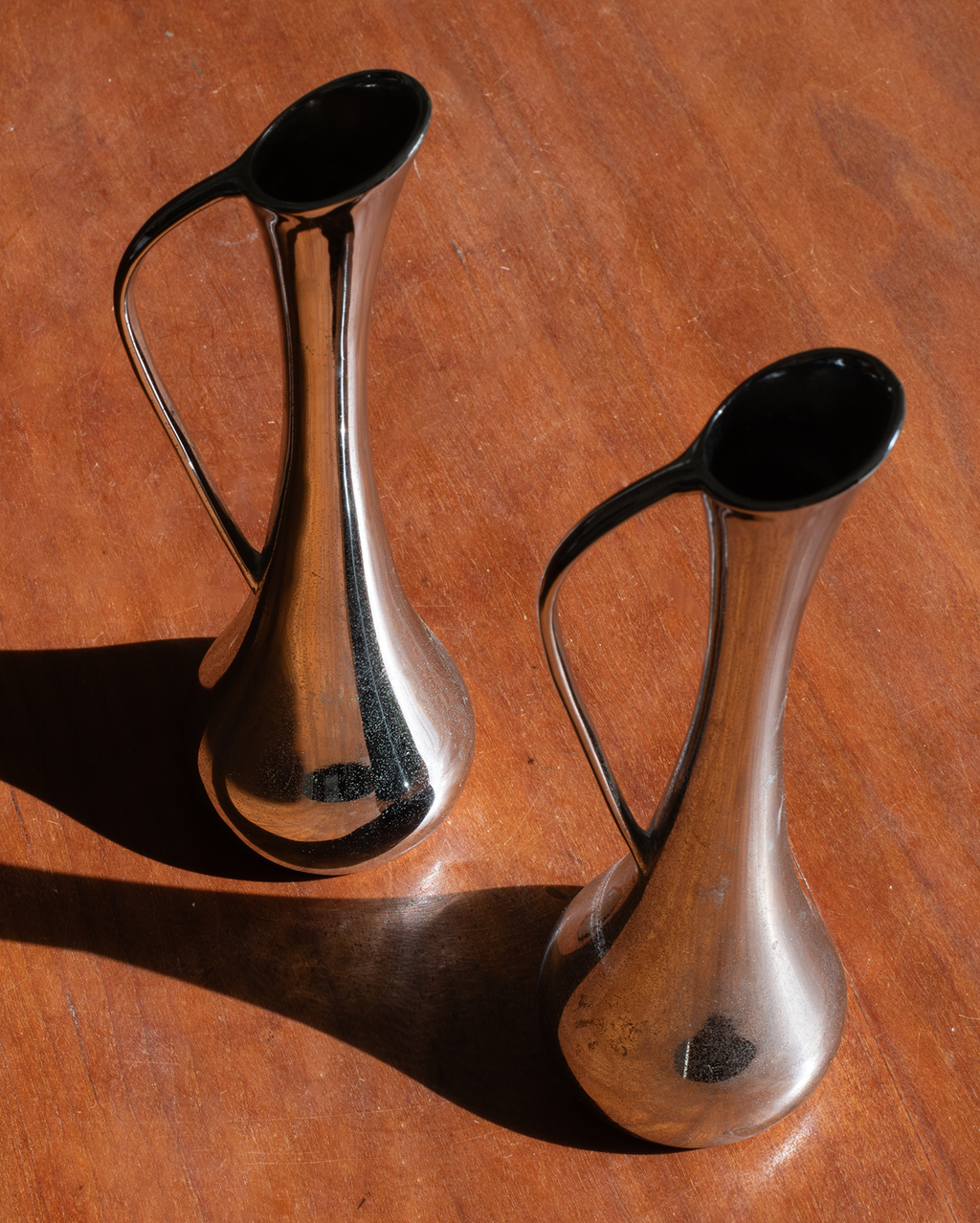 Two polished metal pitcher-like vessels with curved handles and flared rims, photographed on a warm brown wooden surface with strong sunlight and shadows.