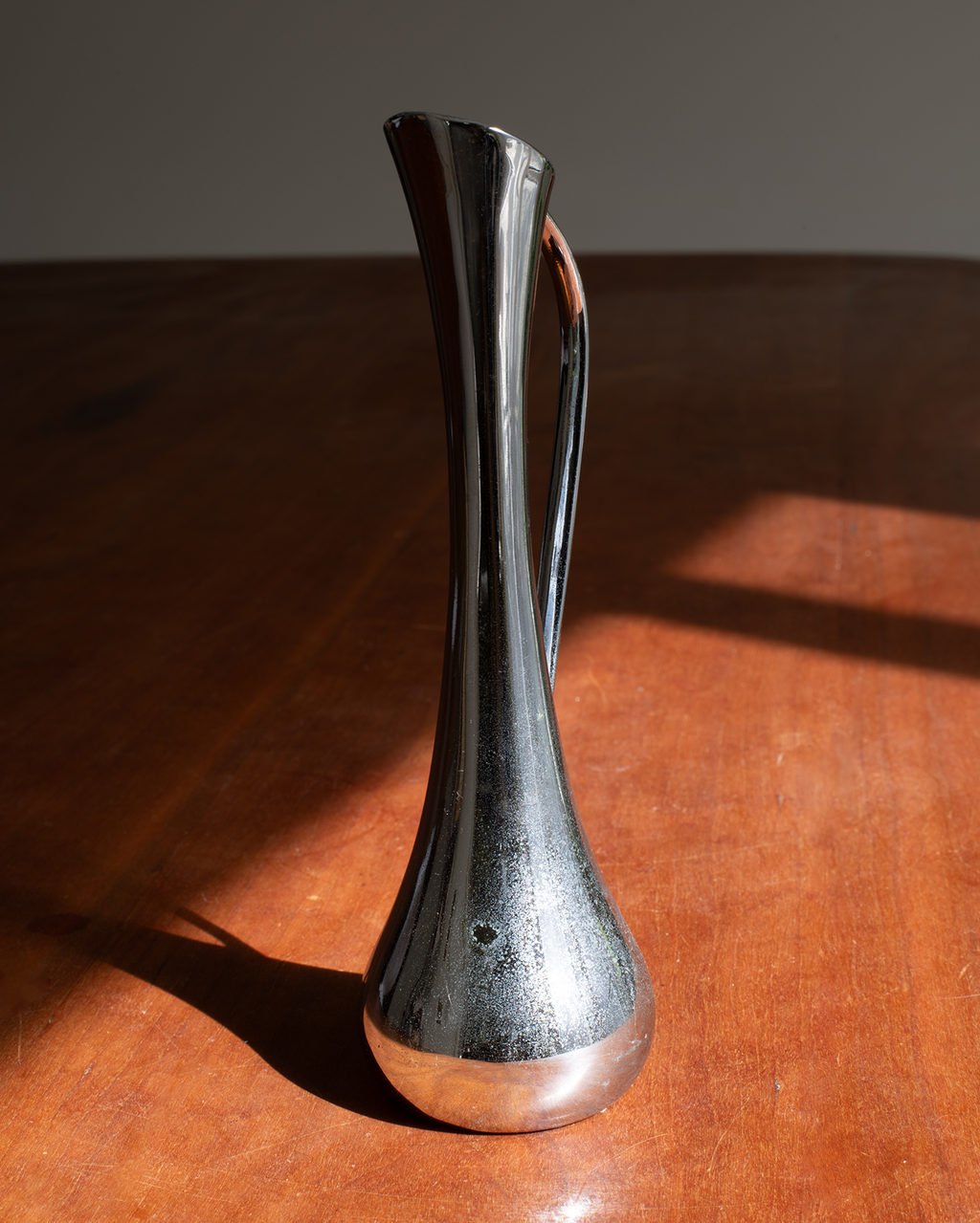 A modern sculptural metallic vase with a flared neck and curved handle, standing on a polished wooden table under direct sunlight.