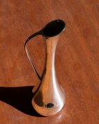 A small polished wooden pitcher with a dark handle and black rim, photographed on a warm brown wooden tabletop with a strong shadow.