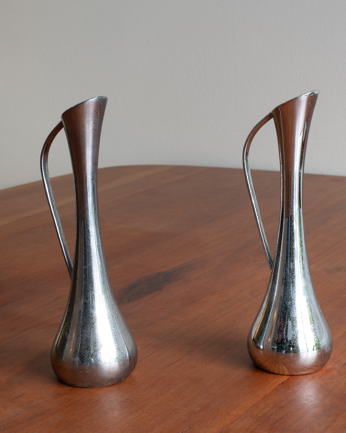 Two vintage polished silver metal pitchers with curved handles, displayed side-by-side on a wooden table against a neutral background.