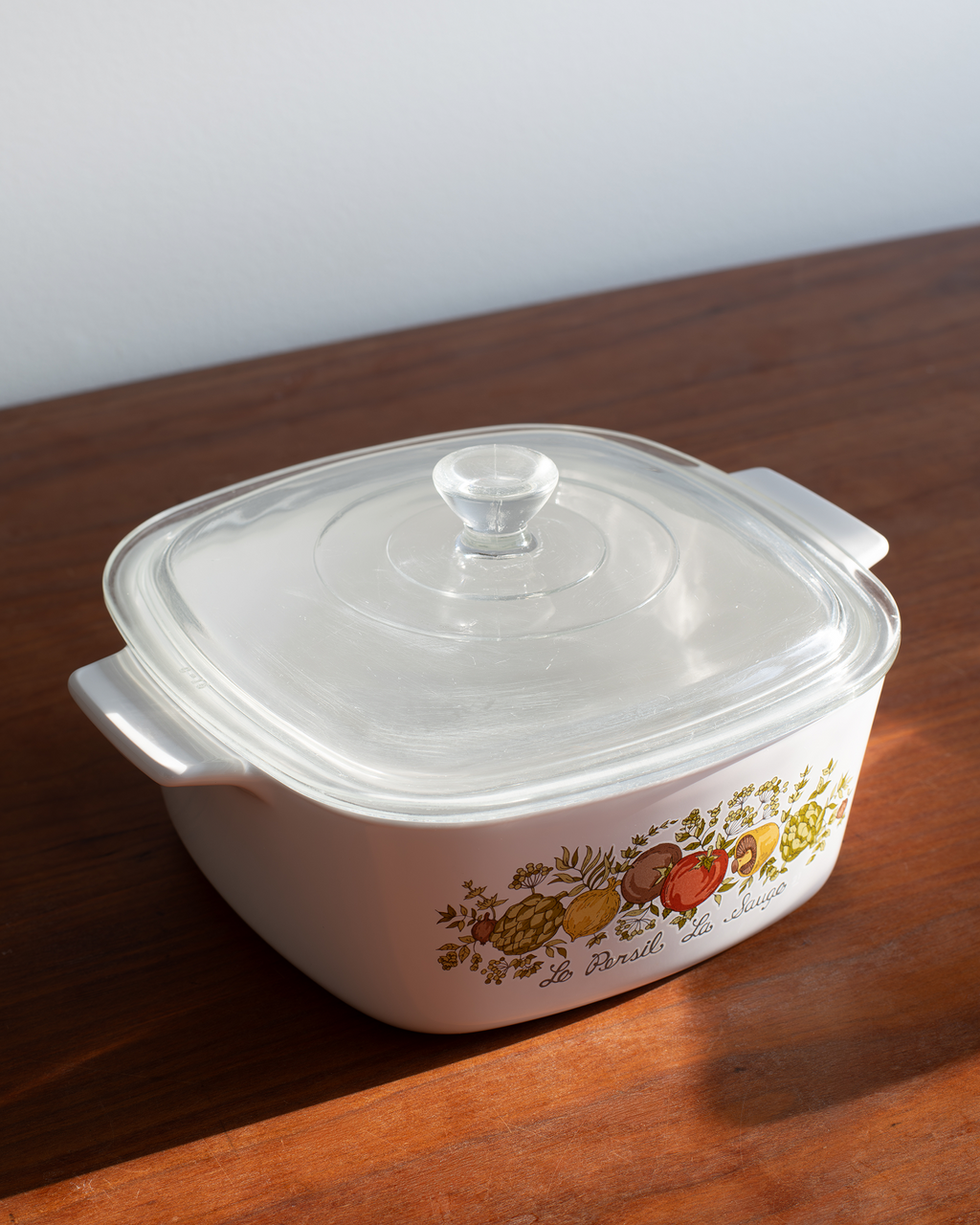 A white rectangular glass casserole dish with a clear lid and side handles, decorated with a colorful fruit pattern, resting on a wooden table.