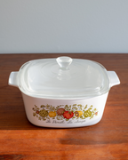 A white glass casserole dish with a matching glass lid sits on a wooden table against a light gray wall. The front of the dish features a colorful fruit illustration.