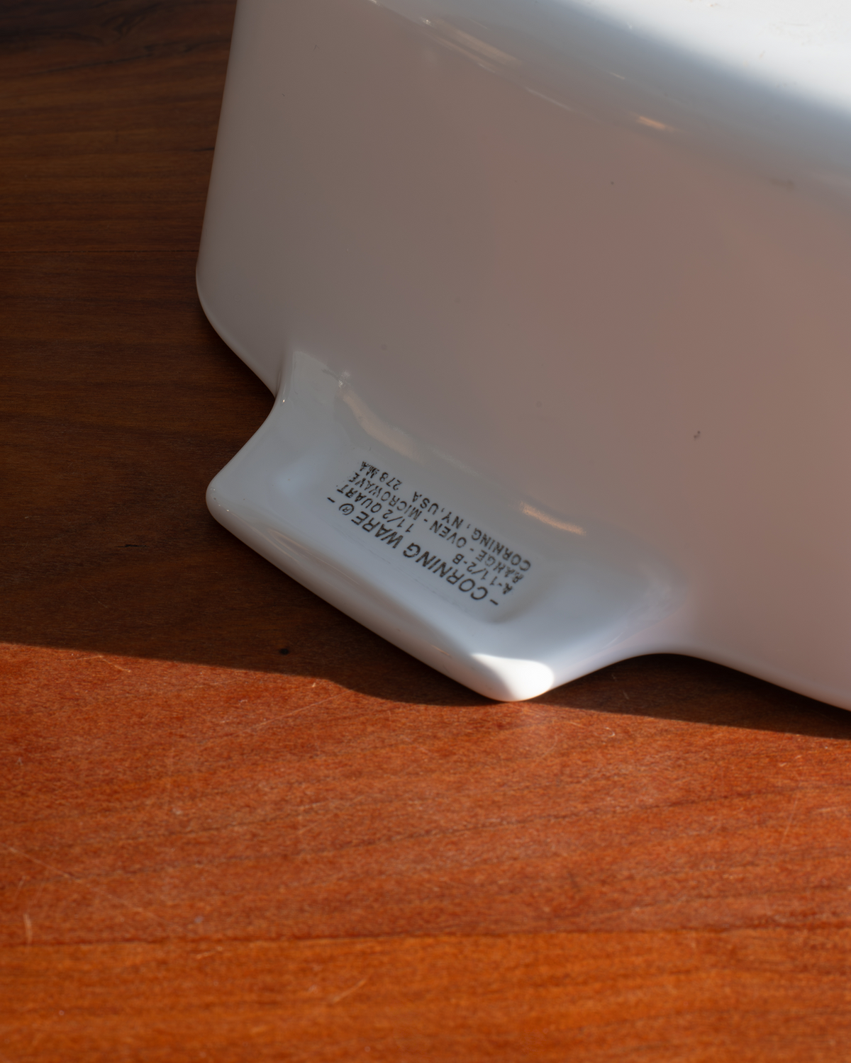 Close-up of the bottom edge of a white ceramic baking dish or casserole dish on a wooden surface, showing printed text including 'CORNING WARE'.