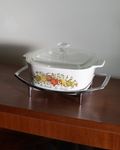 A white ceramic casserole dish with a glass lid, featuring a colorful fruit illustration and the text 'la cuisine de Marie' on the front, displayed on a clear glass stand over a wooden table against a plain wall.