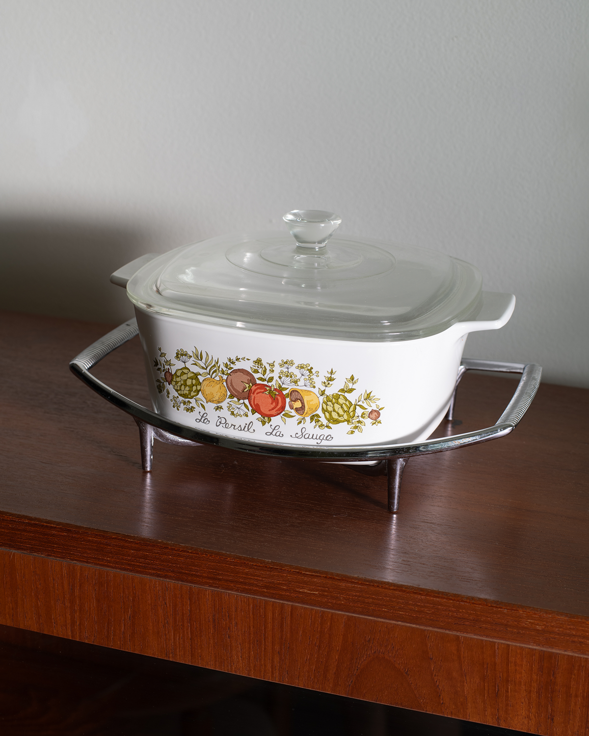 A white ceramic casserole dish with a glass lid, featuring a colorful fruit illustration and the text 'la cuisine de Marie' on the front, displayed on a clear glass stand over a wooden table against a plain wall.