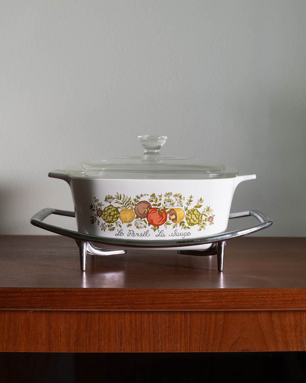 A white ceramic casserole dish with a glass lid, featuring a colorful fruit illustration and the text 'de l'art et du choix' on the side, displayed on a silver metal stand on a wooden table against a gray wall.