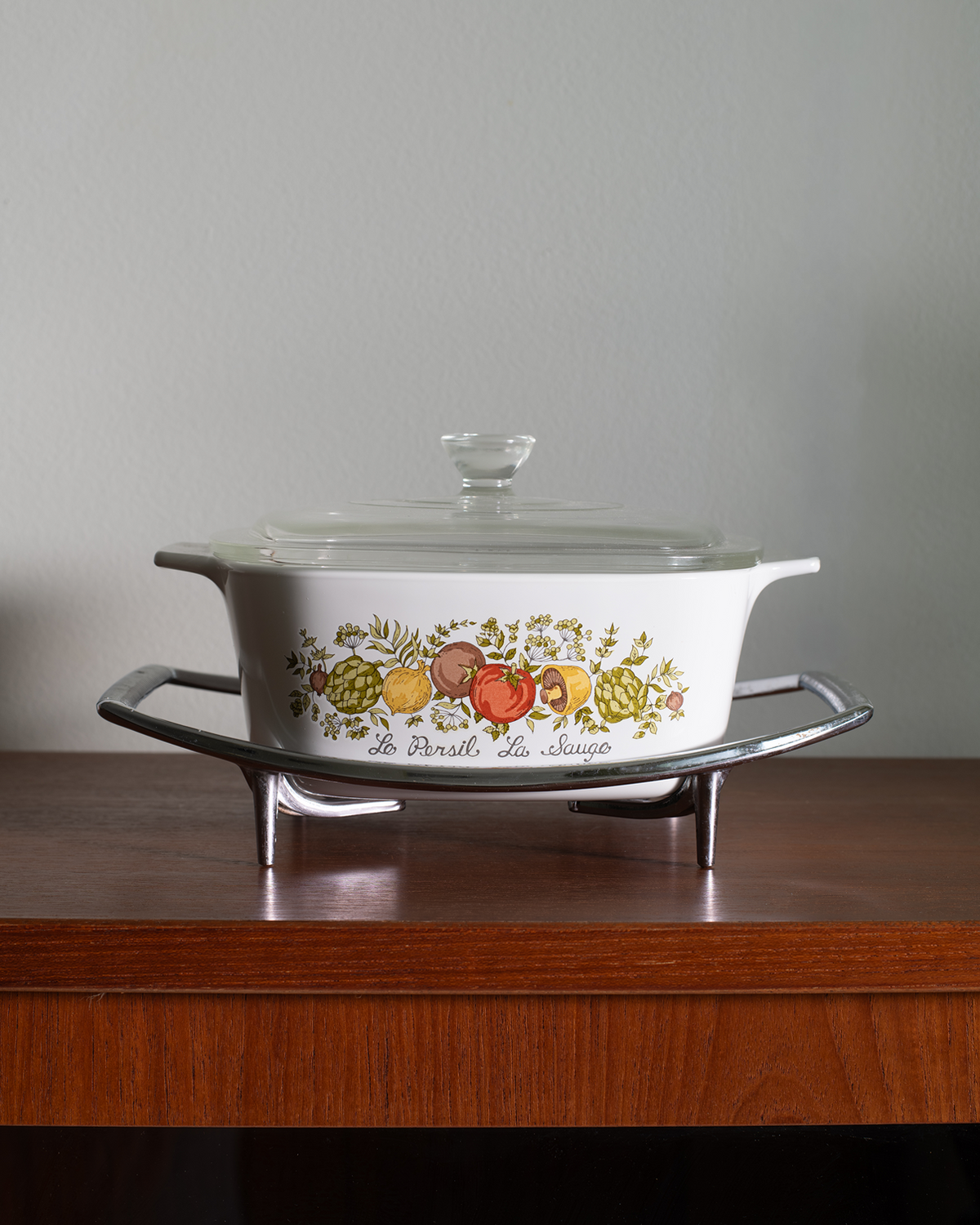 A white ceramic casserole dish with a glass lid, featuring a colorful fruit illustration and the text 'de l'art et du choix' on the side, displayed on a silver metal stand on a wooden table against a gray wall.