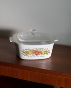 A white glass casserole dish with a clear lid sits on a dark wooden table against a gray wall. The front of the dish is decorated with a colorful fruit illustration and the text 'de Provence de la cuisine'.