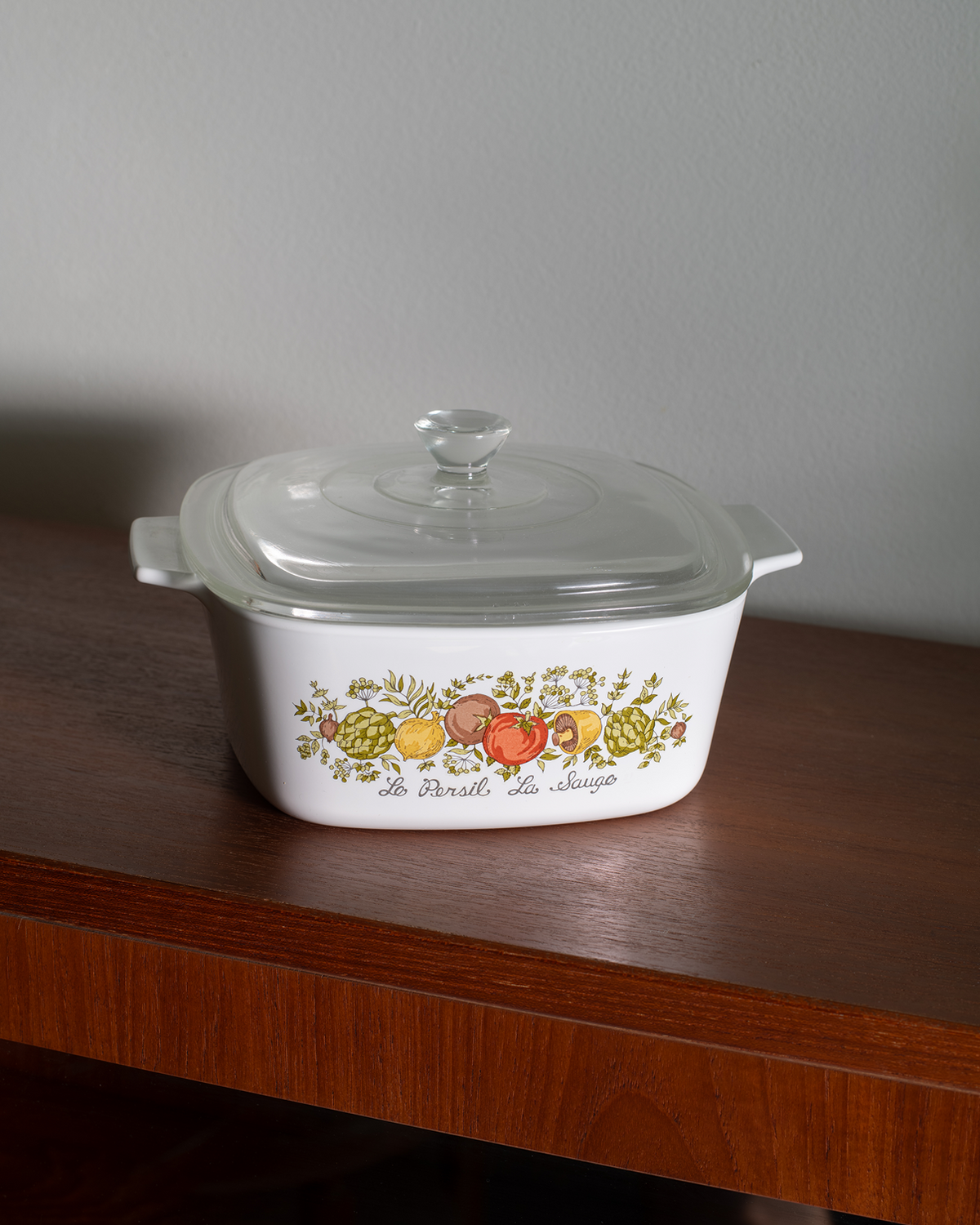 A white glass casserole dish with a clear lid sits on a dark wooden table against a gray wall. The front of the dish is decorated with a colorful fruit illustration and the text 'de Provence de la cuisine'.