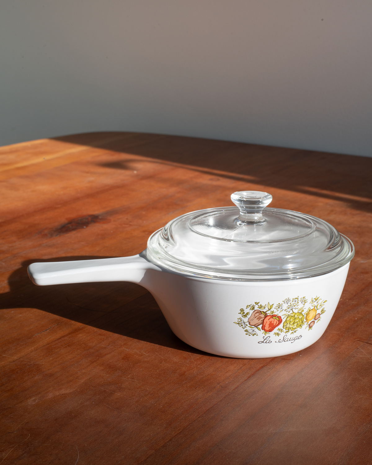 A white La Marécha saucepan with a clear glass lid sits on a warm wooden table under direct sunlight.