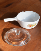 A white ceramic saucepan with a handle and a matching clear glass lid, placed on a wooden tabletop. The saucepan has a small fruit and berry illustration near the base, and the scene is lit by warm natural sunlight with visible shadows.