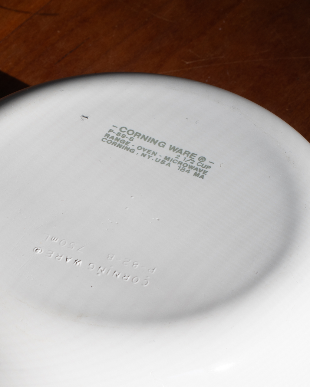 Close-up of the bottom of a white Corning Ware ceramic dish, showing embossed brand text and product information, set against a dark brown wooden surface.