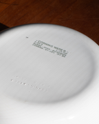 Close-up of the bottom of a white Corning Ware ceramic dish, showing embossed brand text and product information, set against a dark brown wooden surface.