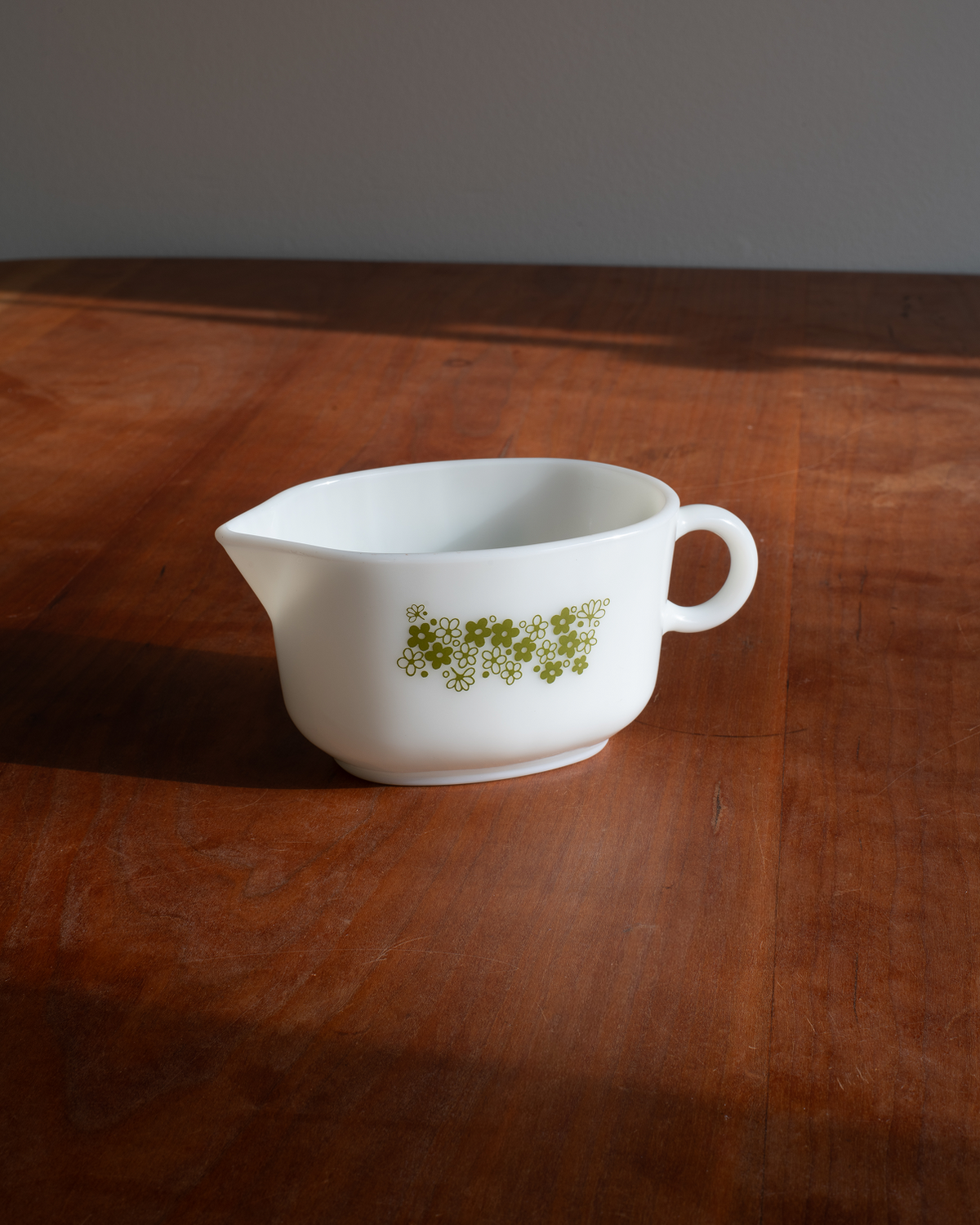 A white ceramic gravy boat with a green floral pattern, sitting on a wooden table under warm, direct sunlight.