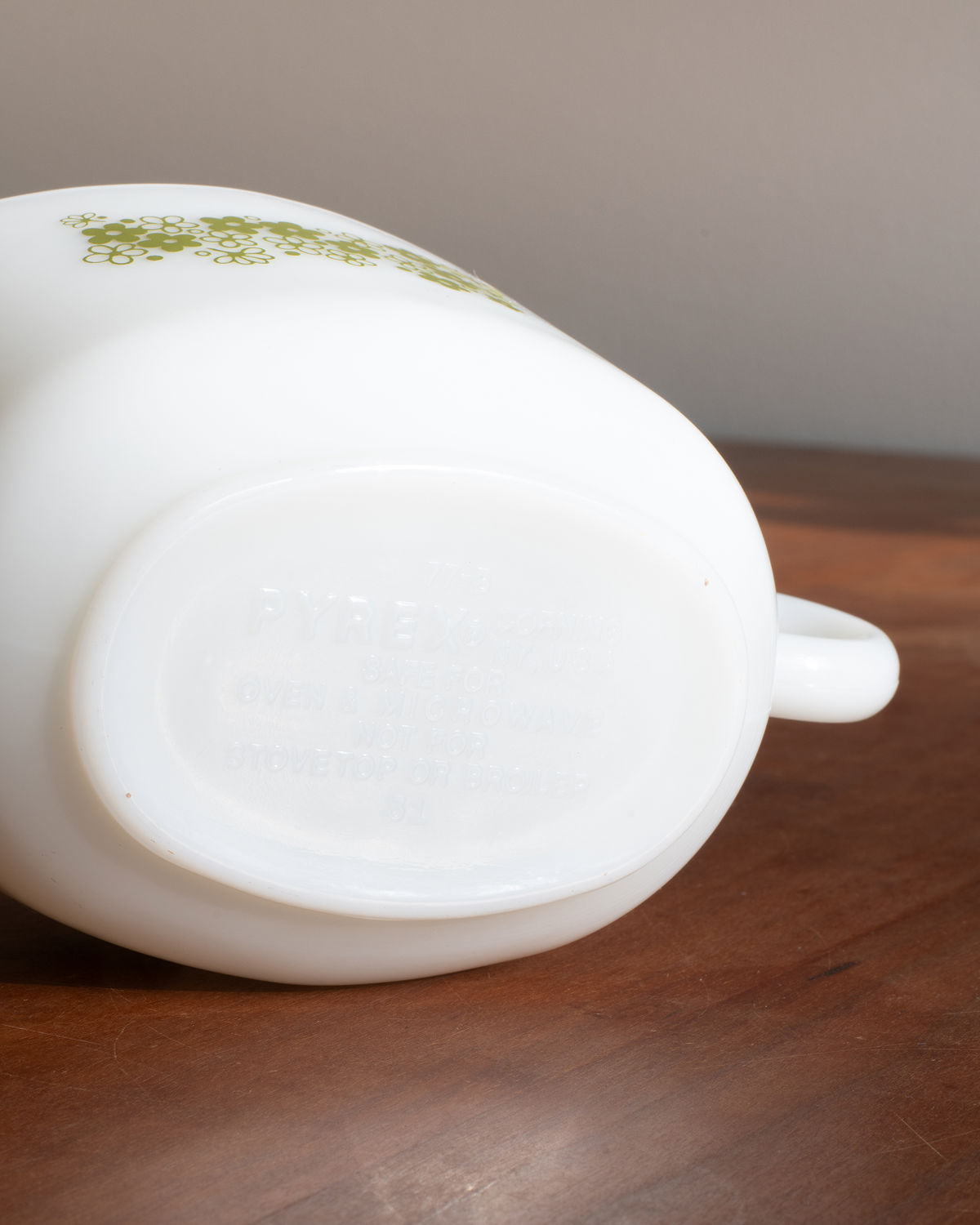 A close-up shot of a vintage white glass pitcher, showing the bottom base with embossed text and a small green floral pattern near the top, resting on a wooden surface.