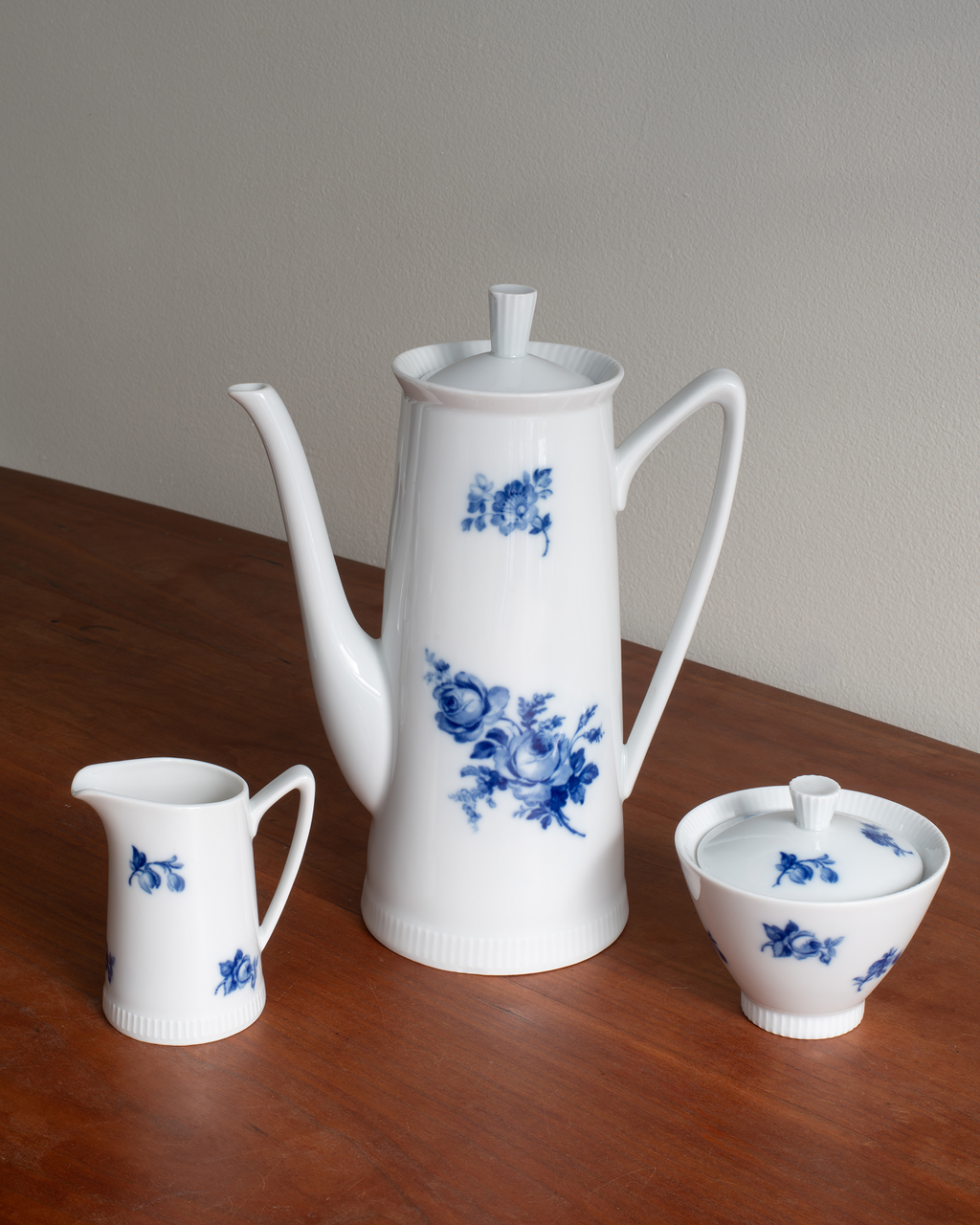 A white porcelain coffee set with blue floral patterns, including a tall coffee pot, a small creamer, and a lidded sugar bowl, arranged on a wooden table against a plain wall.