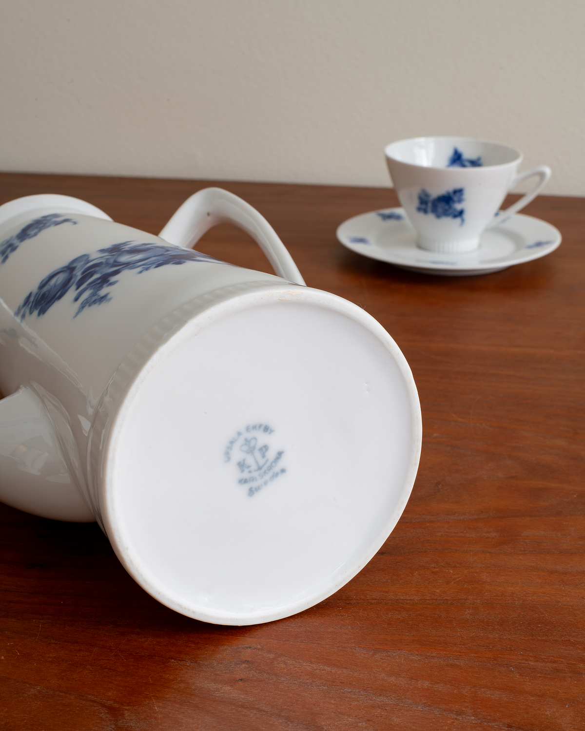 A white ceramic pitcher with blue floral patterns lies on its side on a wooden table, with a matching white teacup and saucer in the background.