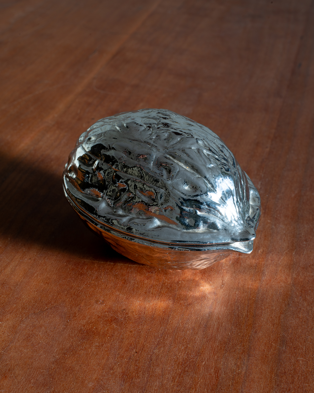 A polished silver metal walnut-shaped decorative object or container, resting on a warm brown wooden surface.